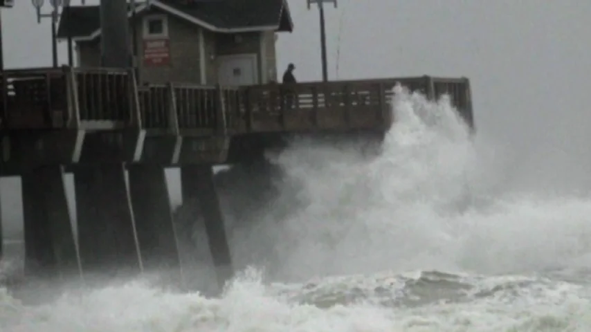 Storm waves breaking against the pilings of a fishing pier, with a figure on the deck above.